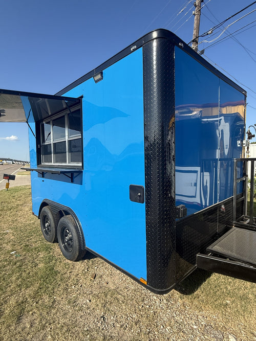 A bright blue enclosed food truck trailer with black trim, a diamond-plate side panel, a service window, and a rear ramp, photographed outdoors on grassy ground under a blue sky.
