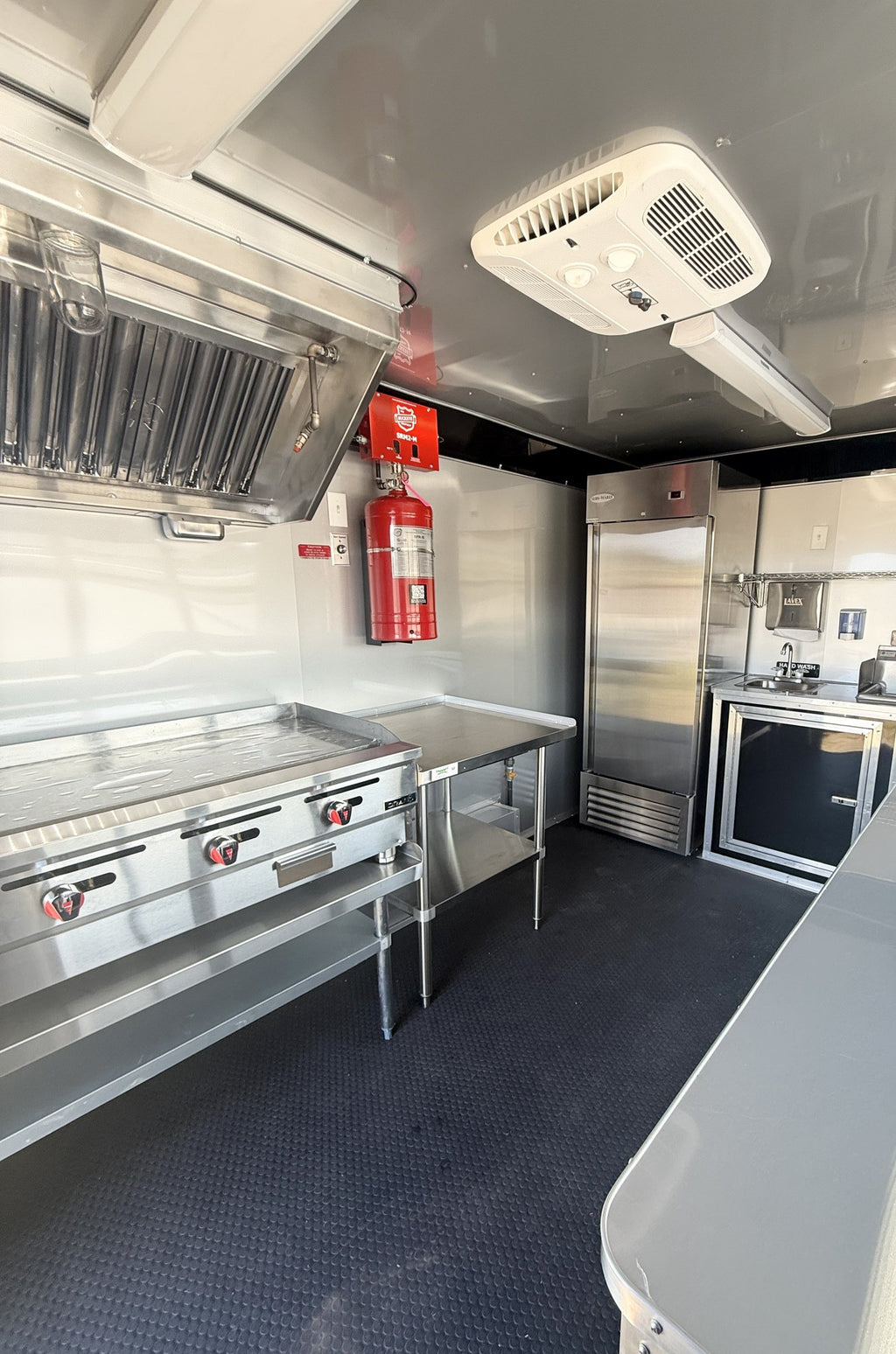 A commercial kitchen interior with stainless steel appliances, including a large range hood, prep table, refrigerator, and small kitchen unit, with a red fire extinguisher mounted on the wall and a dark textured floor.