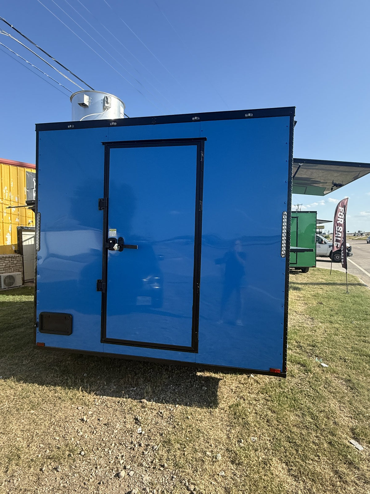 A bright blue portable restroom trailer with a black-framed door, shown outdoors on grass under a blue sky.