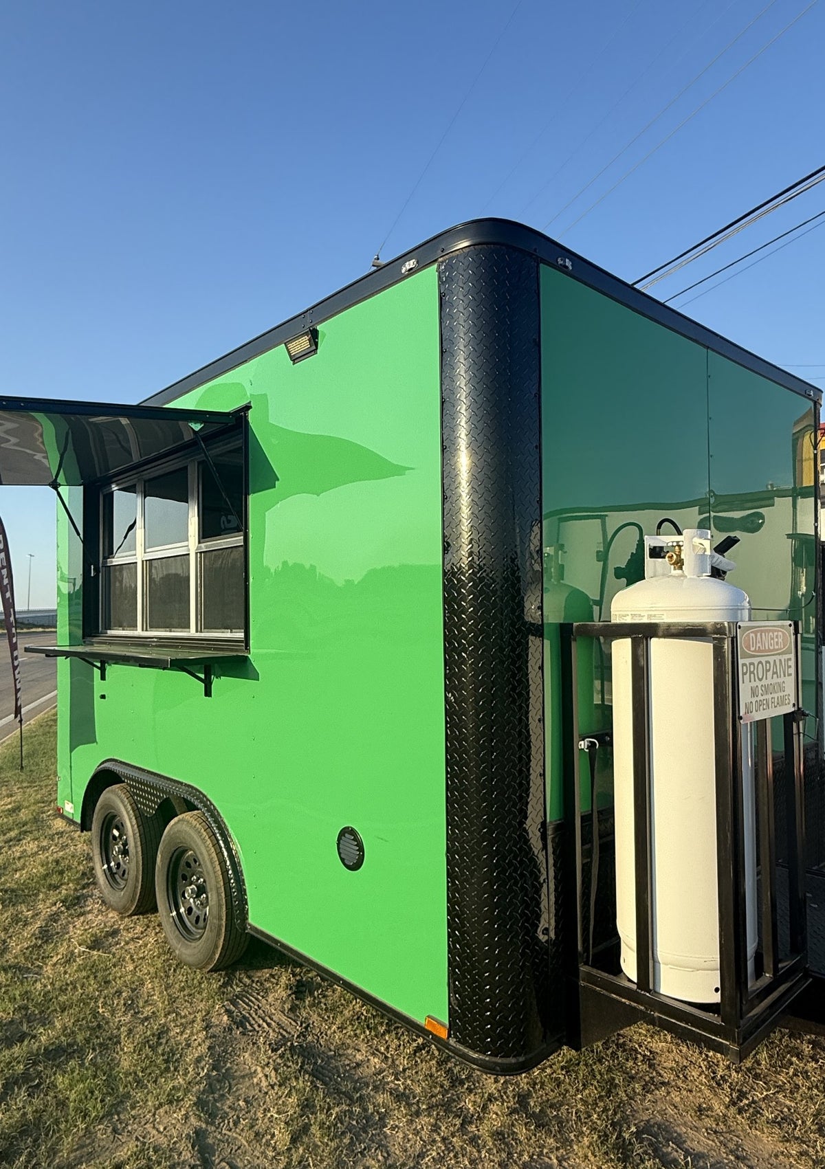 A bright green food truck trailer parked outdoors on grass under a blue sky, with a black diamond-plate bumper, an open service window, and a white propane tank mounted on the side.