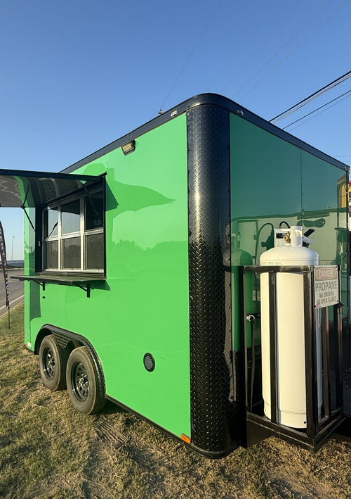 A bright green food truck trailer parked outdoors on grass under a blue sky, with a black diamond-plate bumper, an open service window, and a white propane tank mounted on the side.