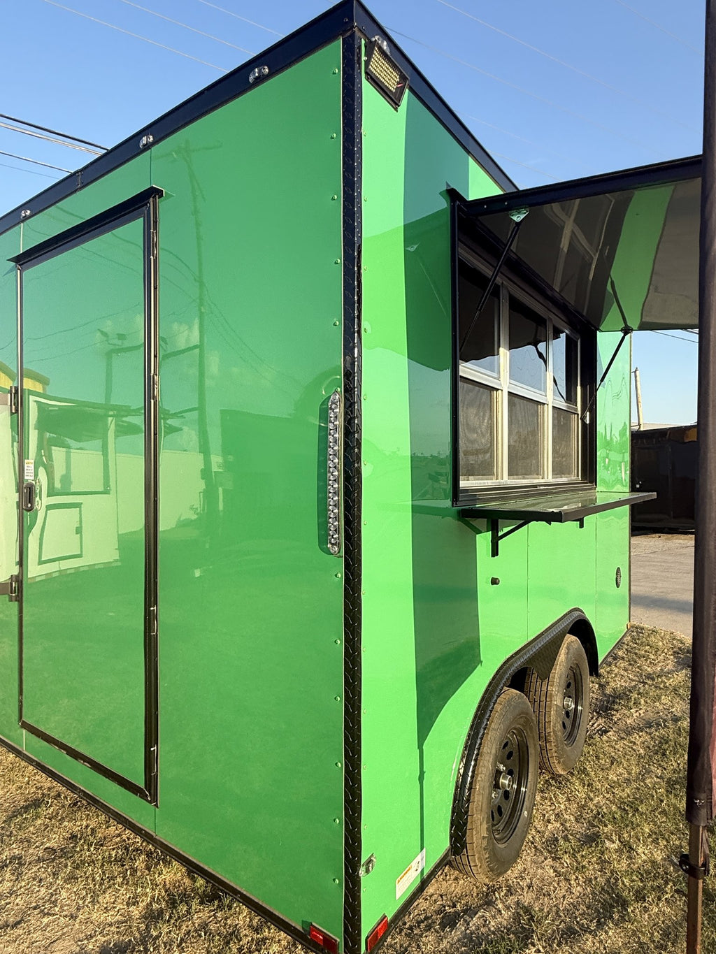 A bright green food truck trailer parked outdoors on dry grass under a blue sky, with a side door and an open serving window visible.