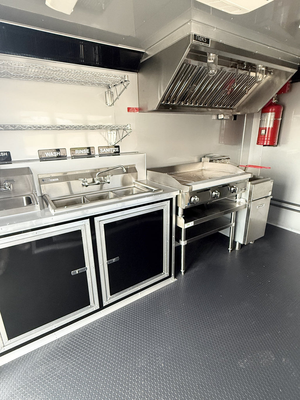 A compact commercial kitchen setup inside a vehicle, featuring stainless steel counters, a sink, a flat-top grill, a large metal range hood, black cabinet doors, a red fire extinguisher, and a textured dark gray floor.