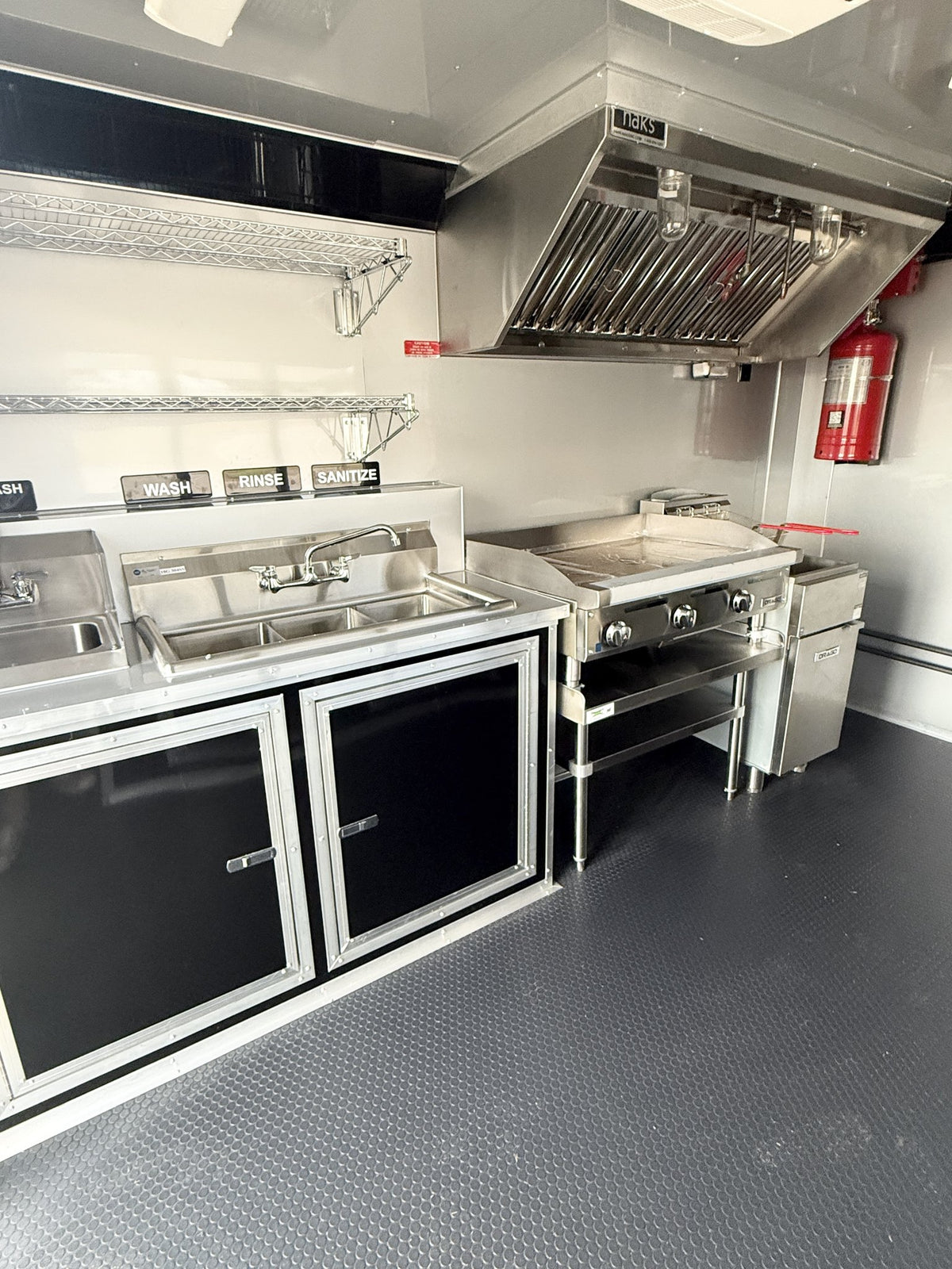 A compact commercial kitchen setup inside a vehicle, featuring stainless steel counters, a sink, a flat-top grill, a large metal range hood, black cabinet doors, a red fire extinguisher, and a textured dark gray floor.