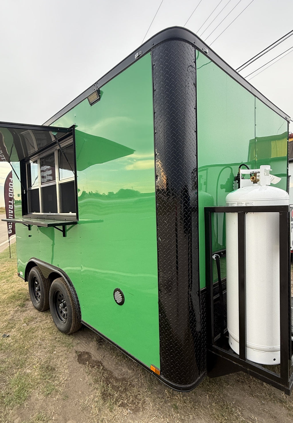 A bright green food truck trailer with black trim, a side service window, and a large white propane tank mounted in a black frame, photographed outdoors on grassy ground under an overcast sky.