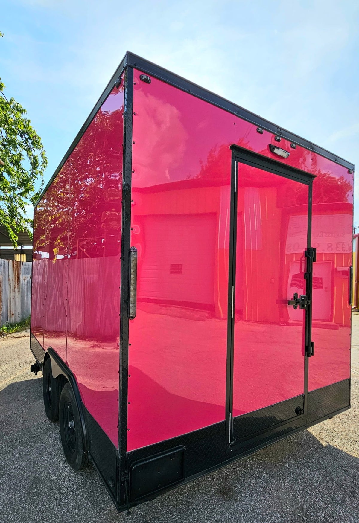 A bright pink enclosed cargo trailer with black trim, side door, and wheels, photographed outdoors on a paved surface under a blue sky.