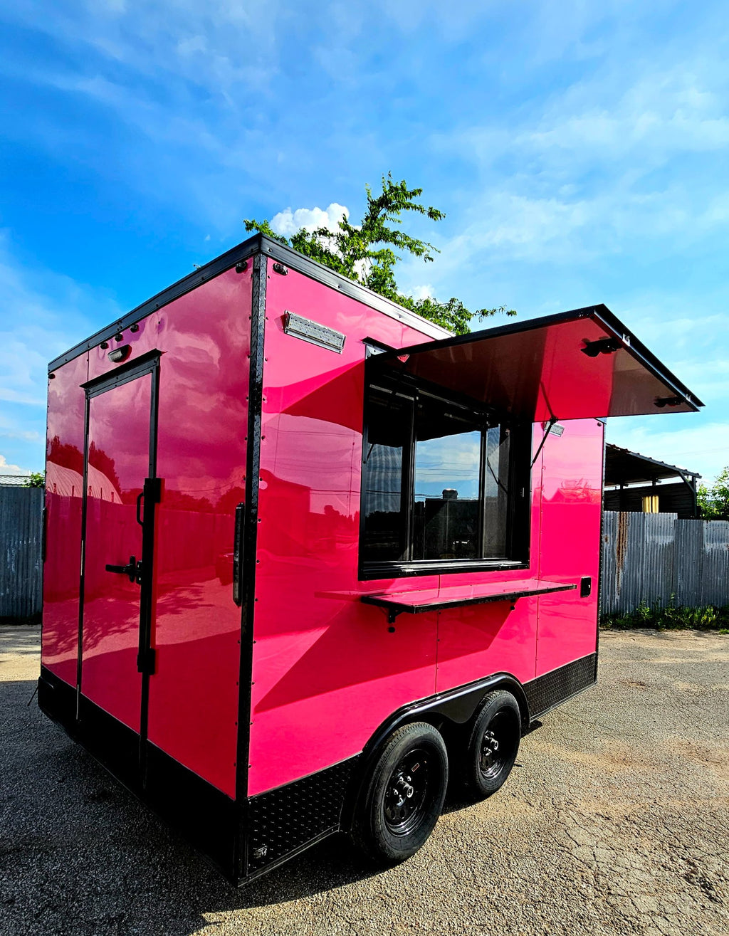 A bright pink food truck trailer with black trim and a raised serving window, parked outdoors on a gravel surface under a blue sky.