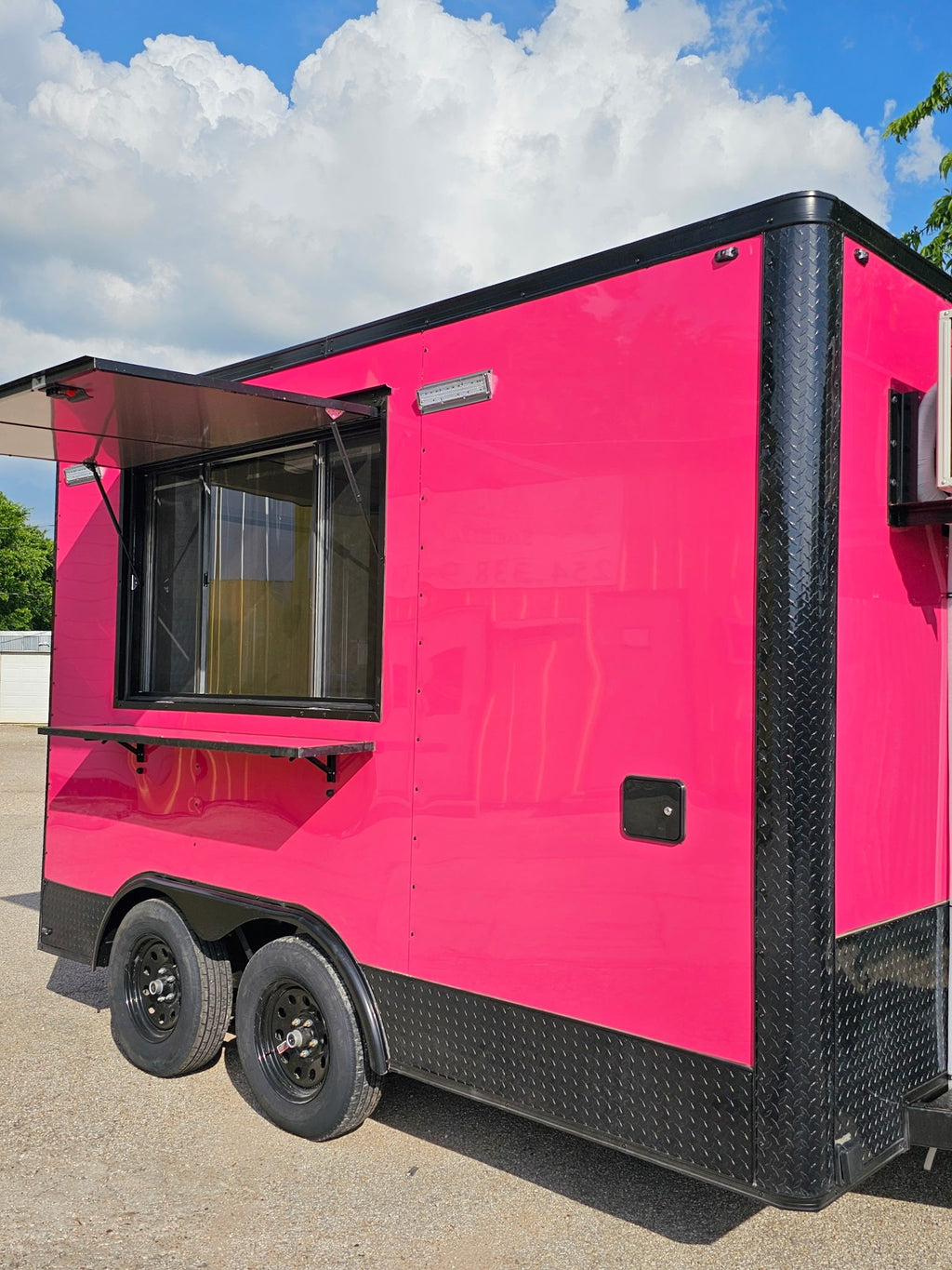 A bright pink food truck trailer with black diamond-plate trim and wheels, parked outdoors on a paved surface under a blue sky with clouds.