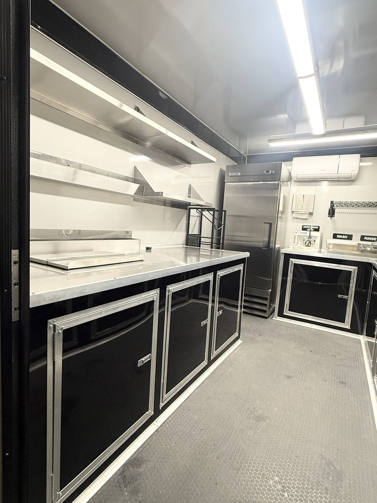A compact mobile kitchen setup inside a trailer, featuring black and stainless steel cabinets, a countertop, a built-in refrigerator, and overhead shelving under bright fluorescent lighting.
