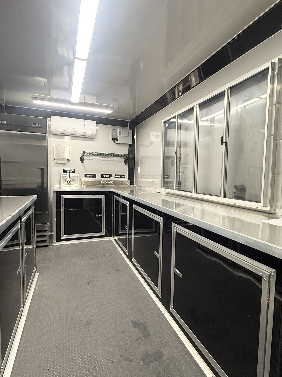 A long, narrow commercial kitchen interior with black cabinets, stainless steel countertops and appliances, large glass service windows, and gray flooring under bright overhead fluorescent lights.
