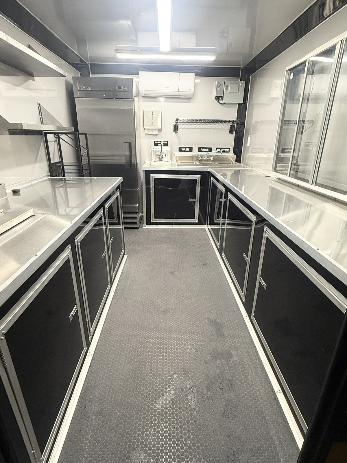 A commercial food prep kitchen interior with stainless steel counters, black cabinets, a sink area, prep table, refrigerator, and gray diamond-plate floor.