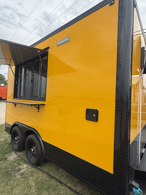 A bright yellow food truck trailer with black diamond-plate trim, a side service window with the awning extended, and dual rear wheels, photographed outdoors on grass under an overcast sky.