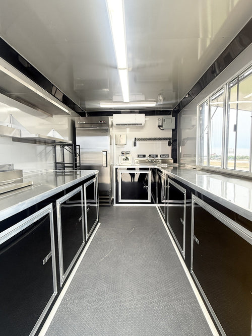 A long, narrow commercial kitchen interior with stainless steel counters, sink, range hood, refrigerator, and sink area, plus gray flooring and large windows on the right side.
