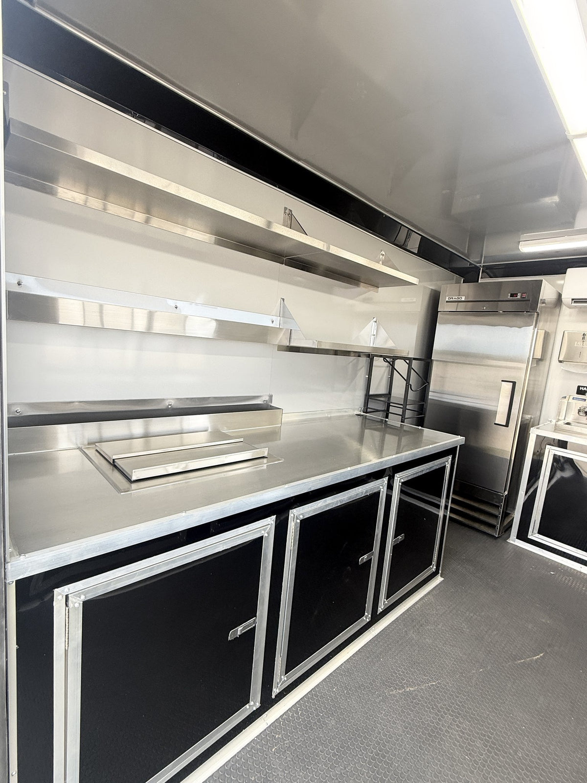 A stainless steel mobile kitchen setup with a workbench, black storage cabinets, shelving, and a large refrigerator inside a vehicle or trailer.