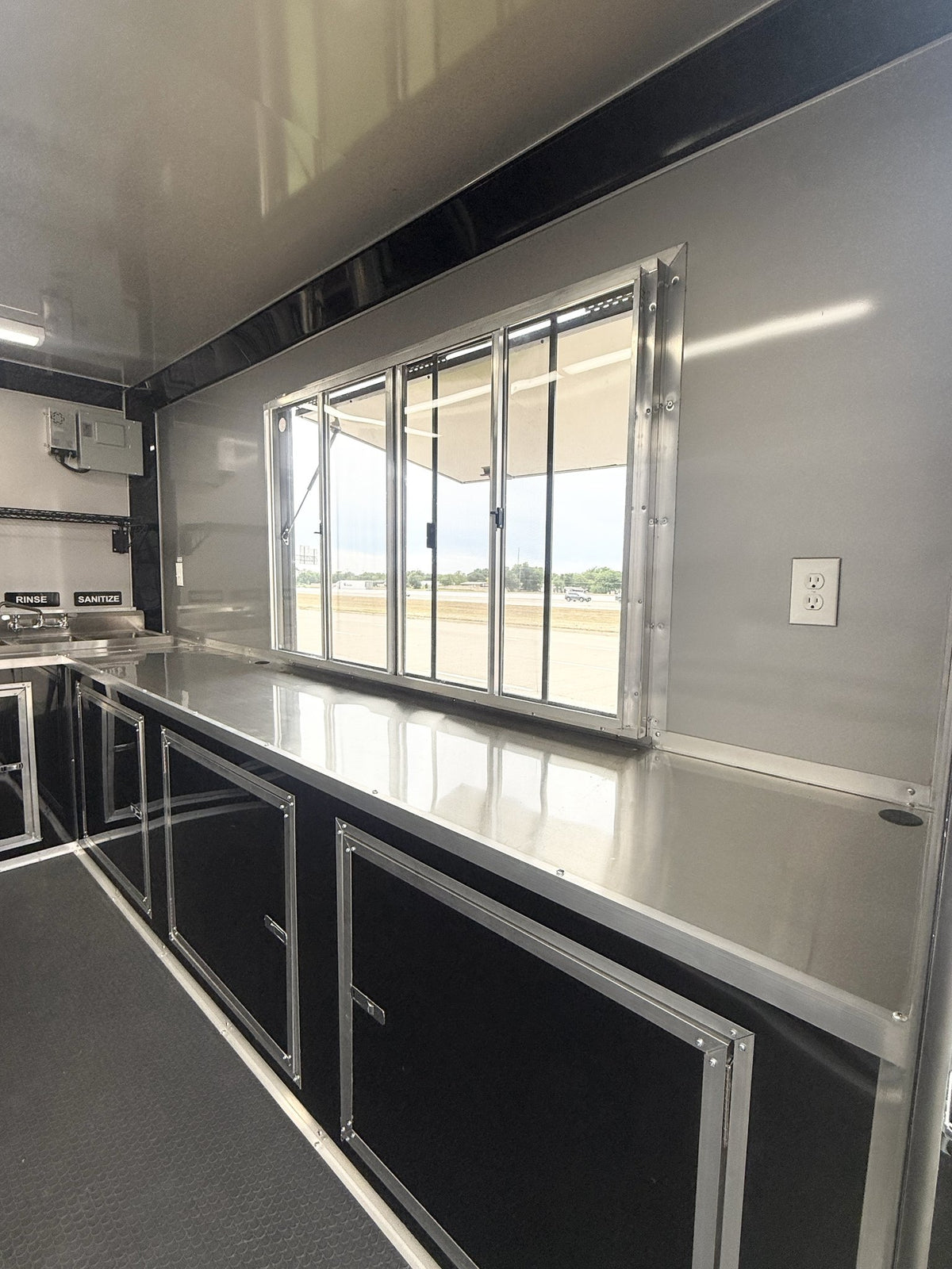 The interior of a trailer or van showing a stainless steel prep counter with a large window, black storage cabinets, and a dark textured floor.