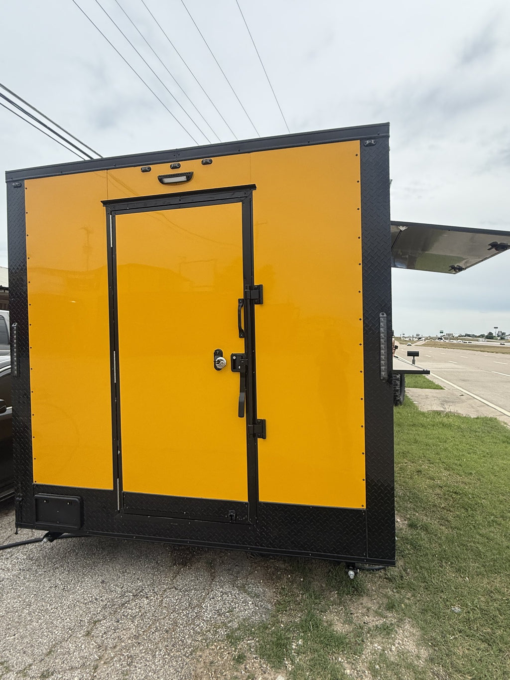 A bright yellow enclosed cargo trailer or utility box with black trim and a single front door, photographed outdoors on gravel beside grass and a paved lot under an overcast sky.