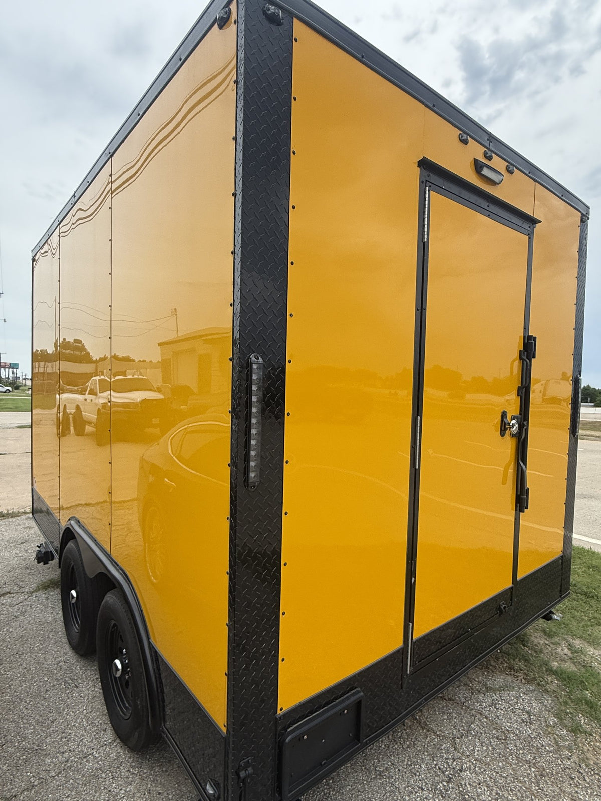 A bright yellow enclosed trailer with black trim, corner bumpers, and a side door, photographed outdoors on a gravel lot under an overcast sky.