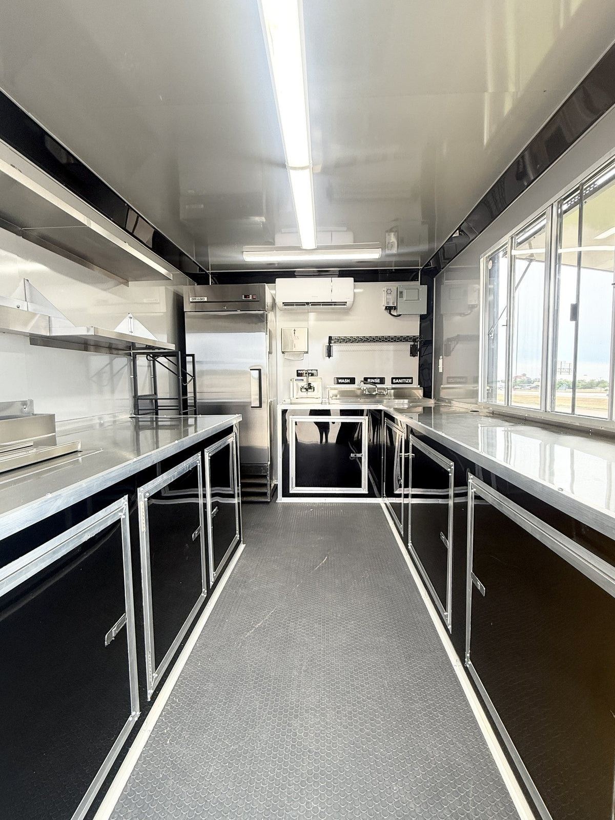 A long, narrow commercial kitchen interior with stainless steel counters, sink, range hood, refrigerator, and sink area, plus gray flooring and large windows on the right side.