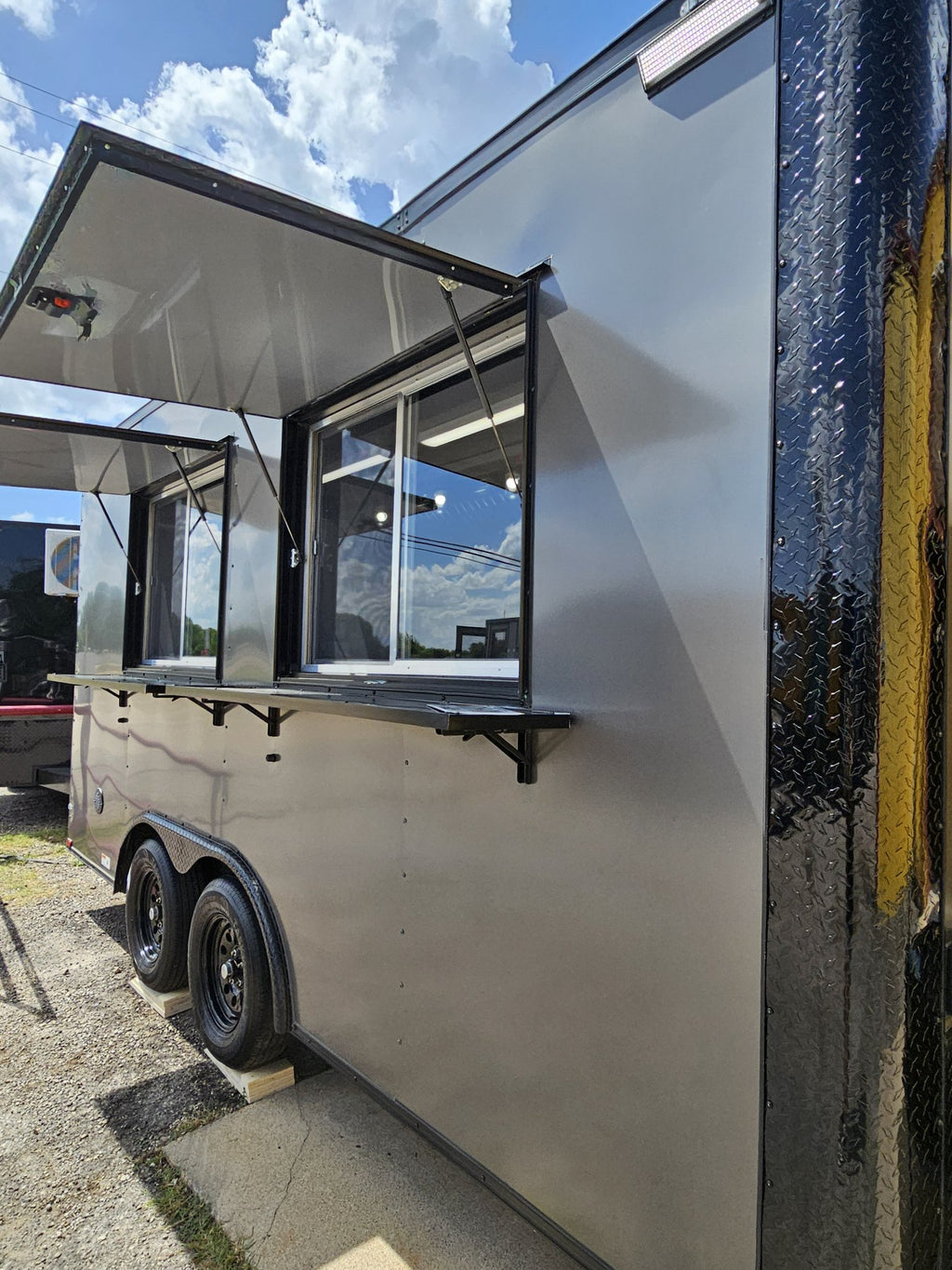 A silver aluminum food truck trailer with black trim, showing an open serving window and awning, parked outdoors on a gravel lot under a blue sky with clouds.