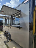A silver aluminum food truck trailer with black trim, showing an open serving window and awning, parked outdoors on a gravel lot under a blue sky with clouds.
