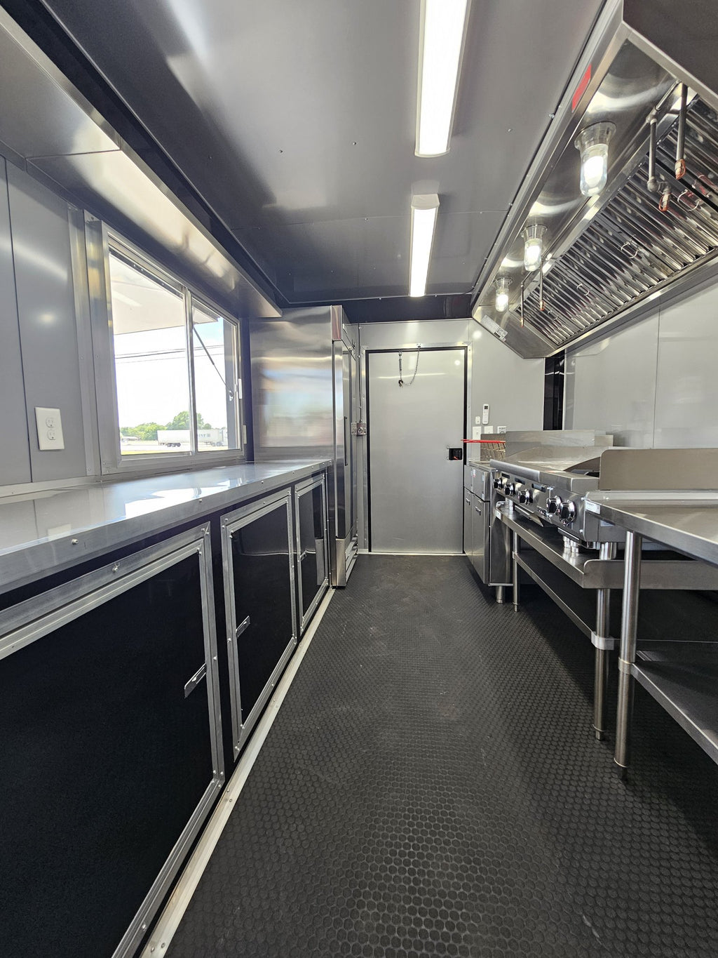 A commercial kitchen interior with stainless steel counters, cabinets, a large range, a ventilation hood, and a dark textured floor.