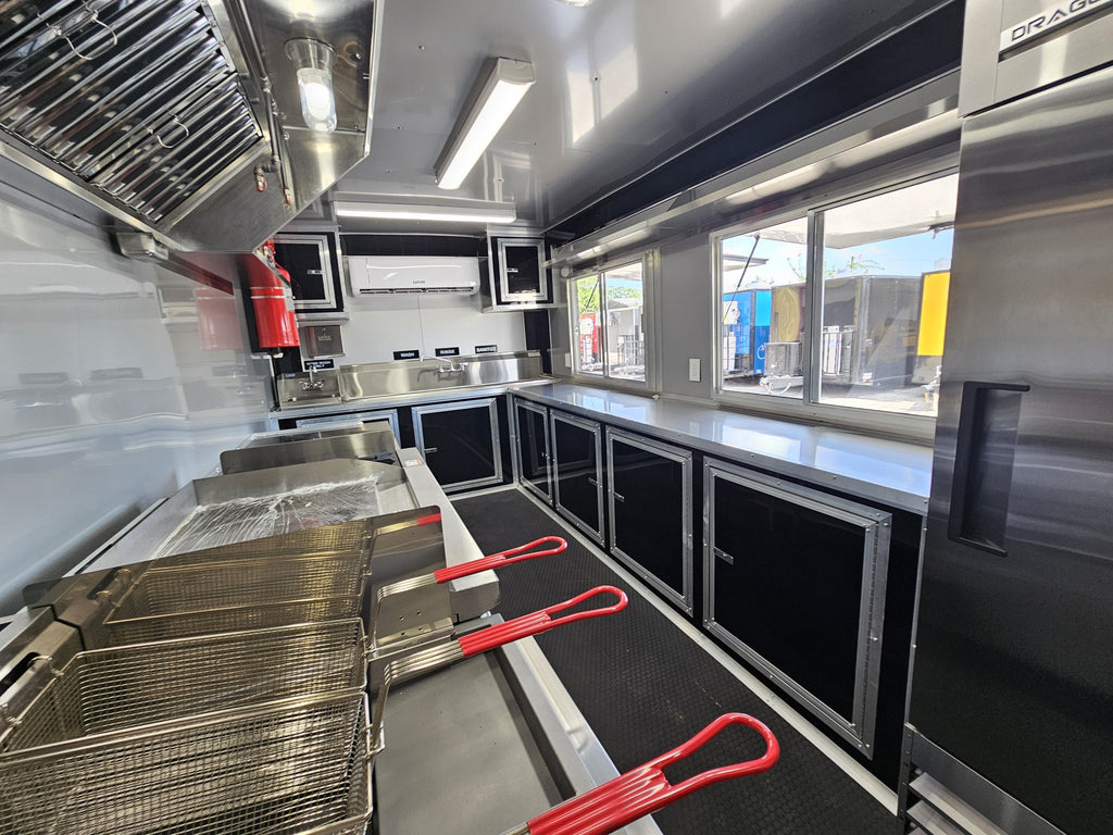 The interior of a stainless steel food truck kitchen, featuring deep fryers, workstations, black storage cabinets, and red handle tools on the floor.