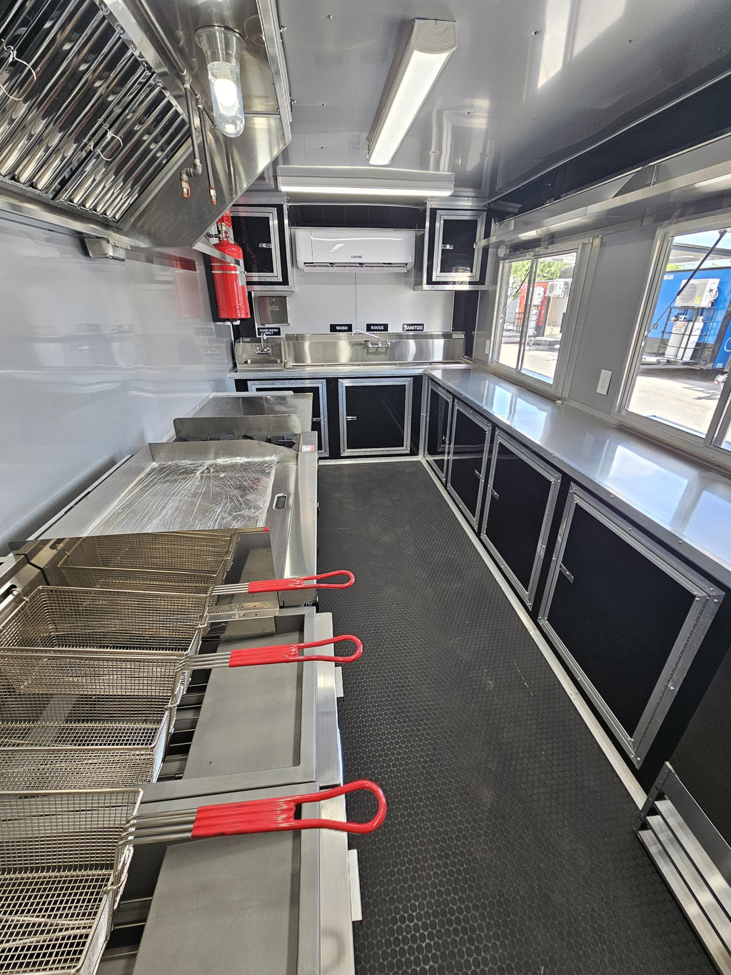 The interior of a mobile kitchen or food truck showing stainless steel cooking equipment, deep fry baskets, black cabinets, a black textured floor, and a bright window on the right side.