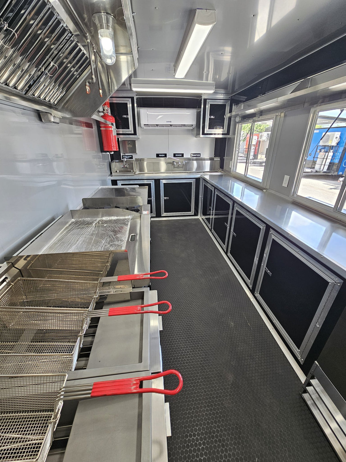 The interior of a mobile kitchen or food truck showing stainless steel cooking equipment, deep fry baskets, black cabinets, a black textured floor, and a bright window on the right side.