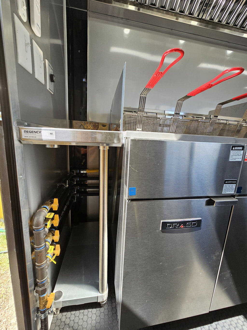 A commercial-grade stainless steel gas range with two deep fryers and red handles, connected to gas pipes with yellow valves.