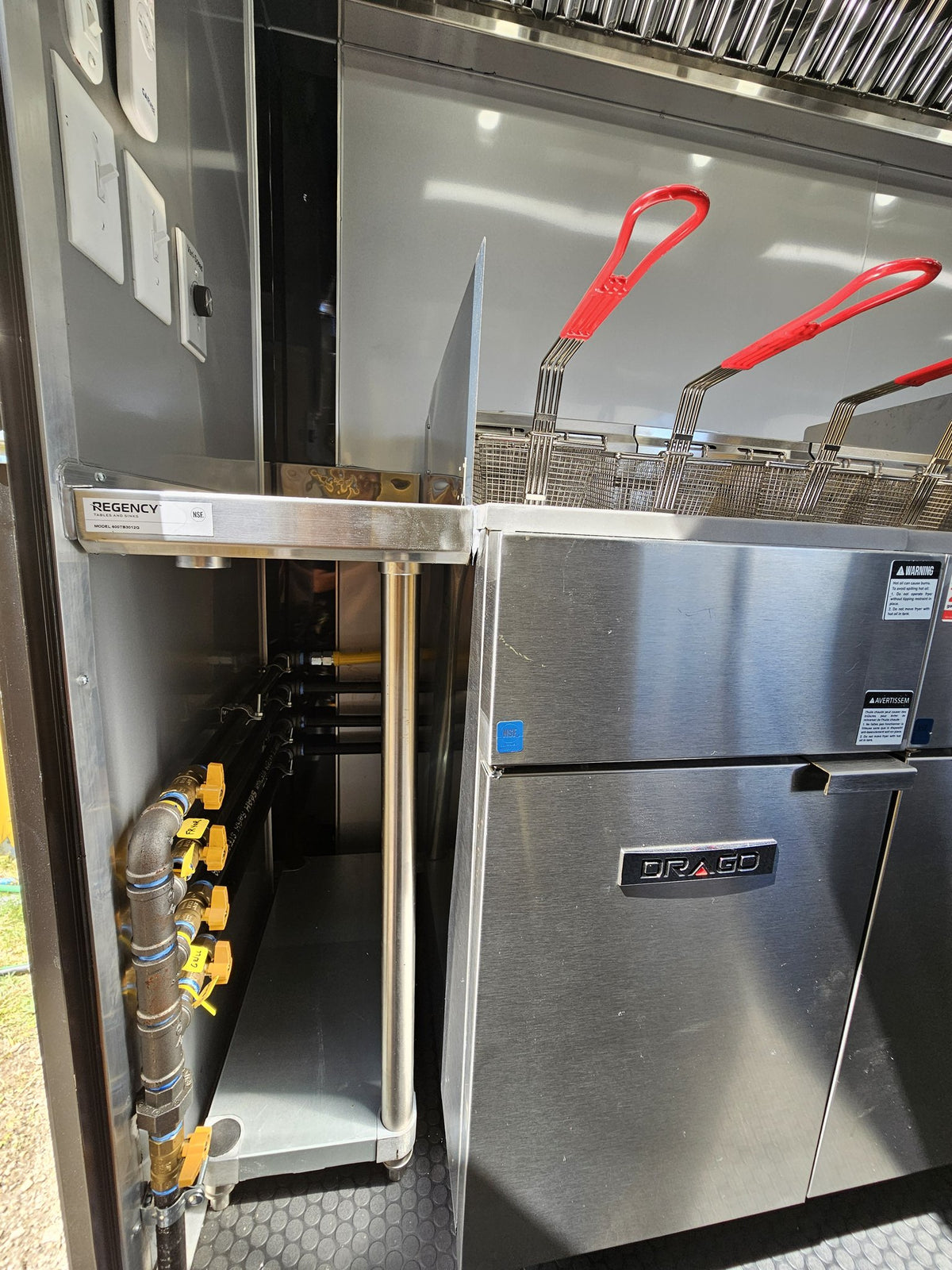 A commercial-grade stainless steel gas range with two deep fryers and red handles, connected to gas pipes with yellow valves.