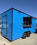 18 FT blue and black cooking trailer parked on gravel under a clear sky, front three-quarter view with the entry door and the serving window awning raised