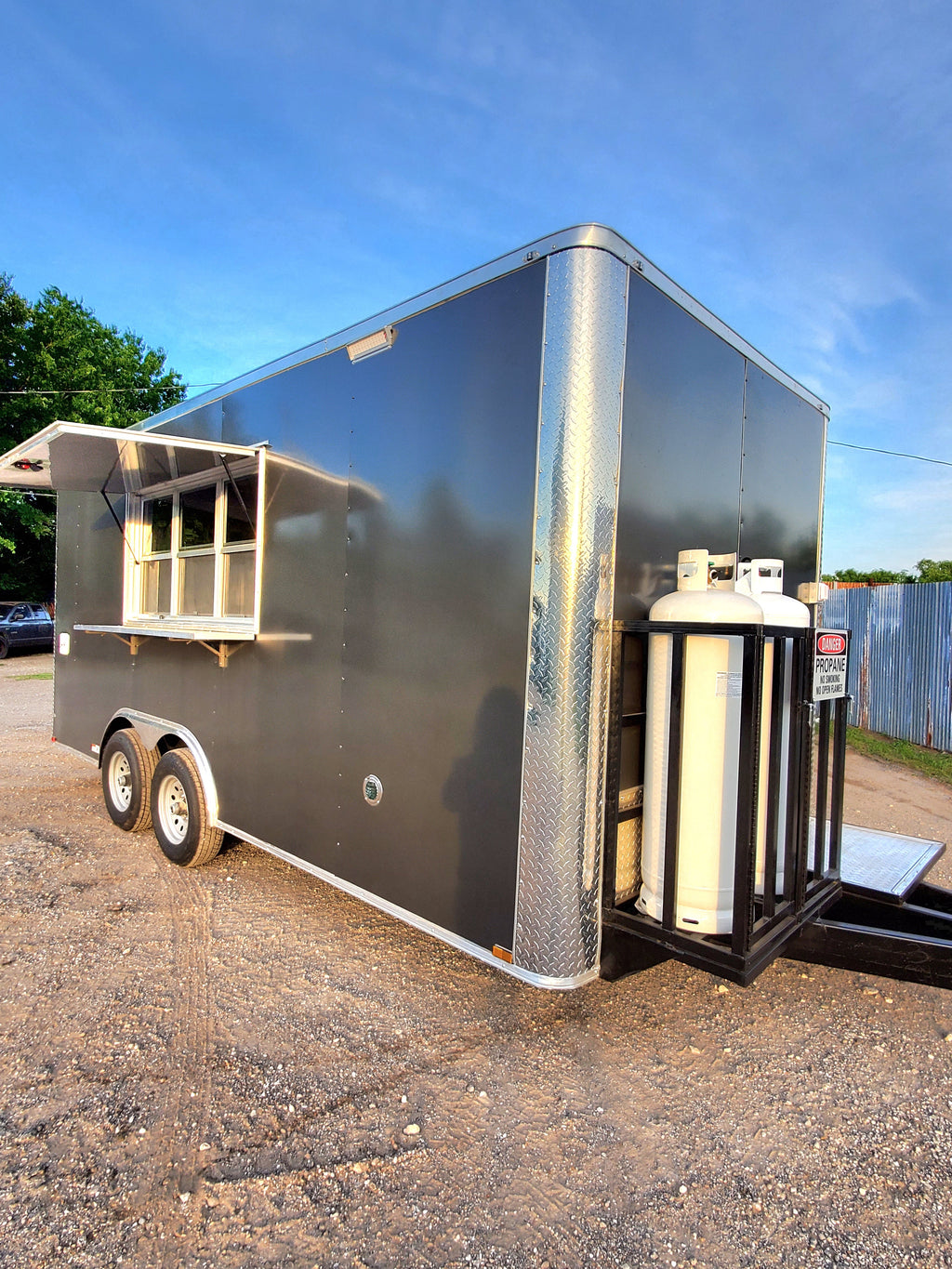 Black food trailer with a silver roof on a gravel surface under a blue sky.