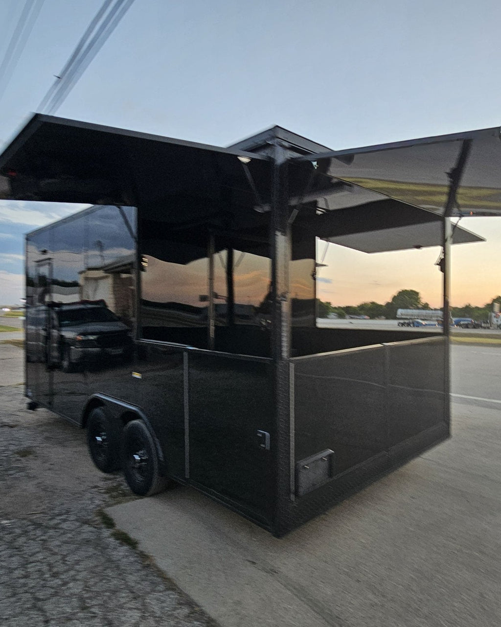 A black commercial food truck trailer with extended side awnings, parked on a paved lot near a road at sunset.