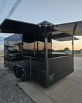 A black commercial food truck trailer with extended side awnings, parked on a paved lot near a road at sunset.
