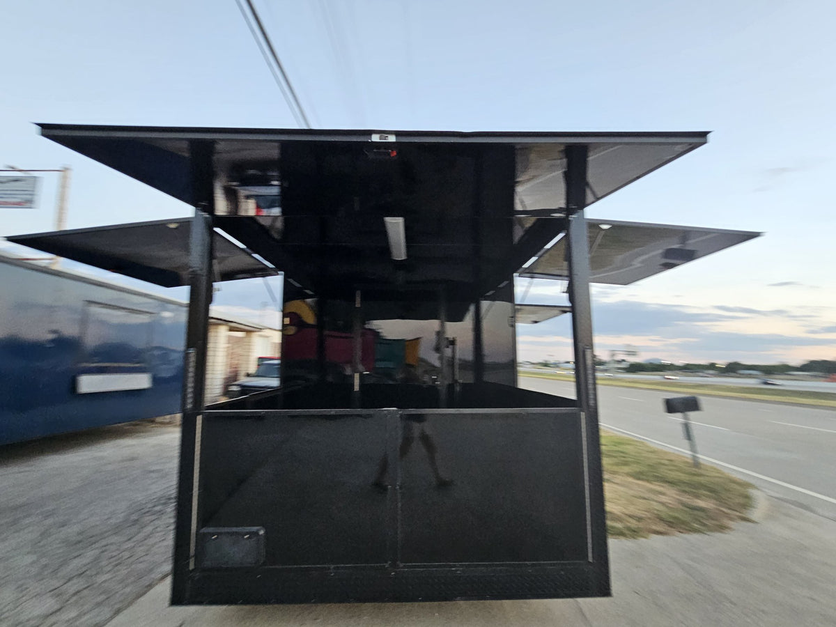 A black metal food truck or kiosk structure photographed outdoors on a paved area beside a road, with its canopy open and side panels extended.