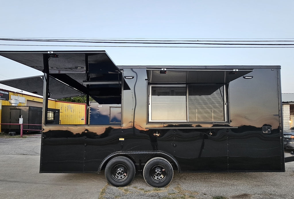 A black metal food truck with extended service windows and awning, parked outdoors on a paved lot with buildings visible in the background.