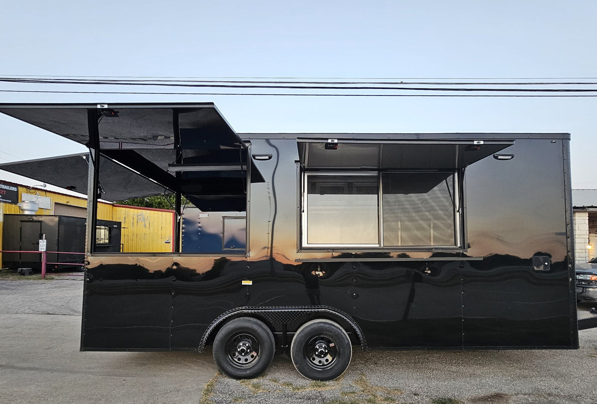 A black metal food truck with extended service windows and awning, parked outdoors on a paved lot with buildings visible in the background.