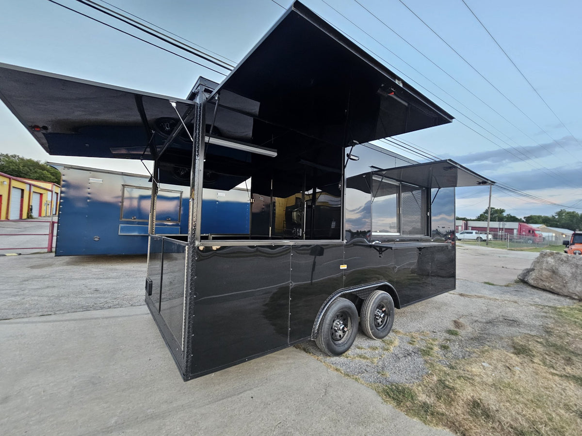 A black commercial food truck trailer with extended service windows and awnings, parked outdoors on a concrete lot with other trailers and utility poles in the background.