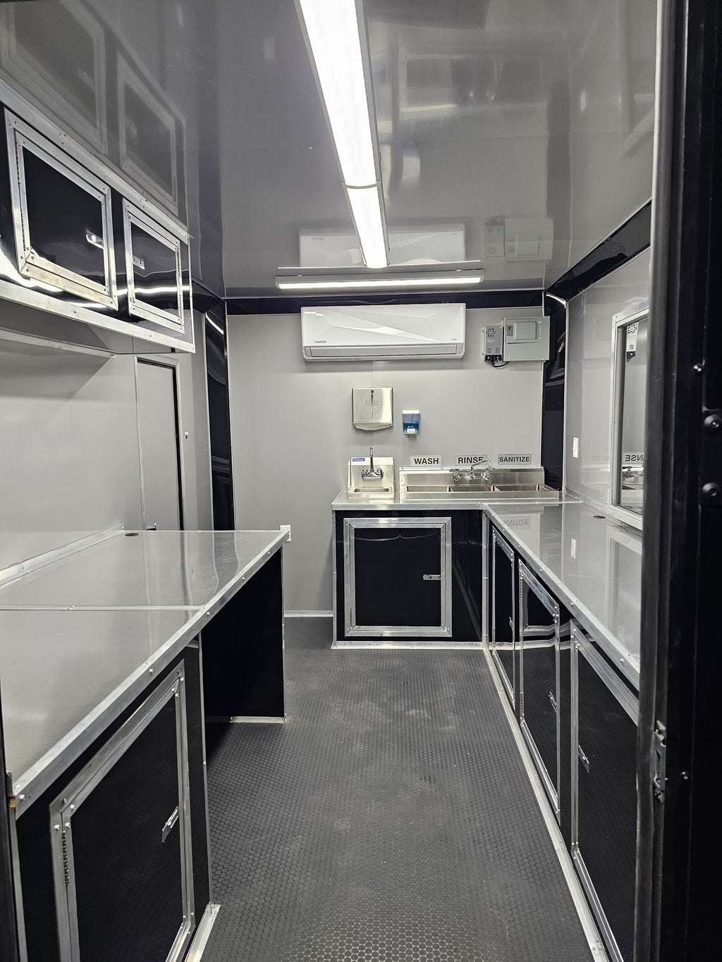 The interior of a customized travel kitchen or mobile prep facility, featuring stainless steel work surfaces, black cabinets, a sink area, and an air conditioner mounted on the back wall.