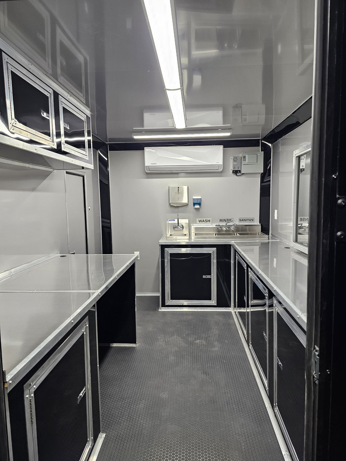 The interior of a customized travel kitchen or mobile prep facility, featuring stainless steel work surfaces, black cabinets, a sink area, and an air conditioner mounted on the back wall.