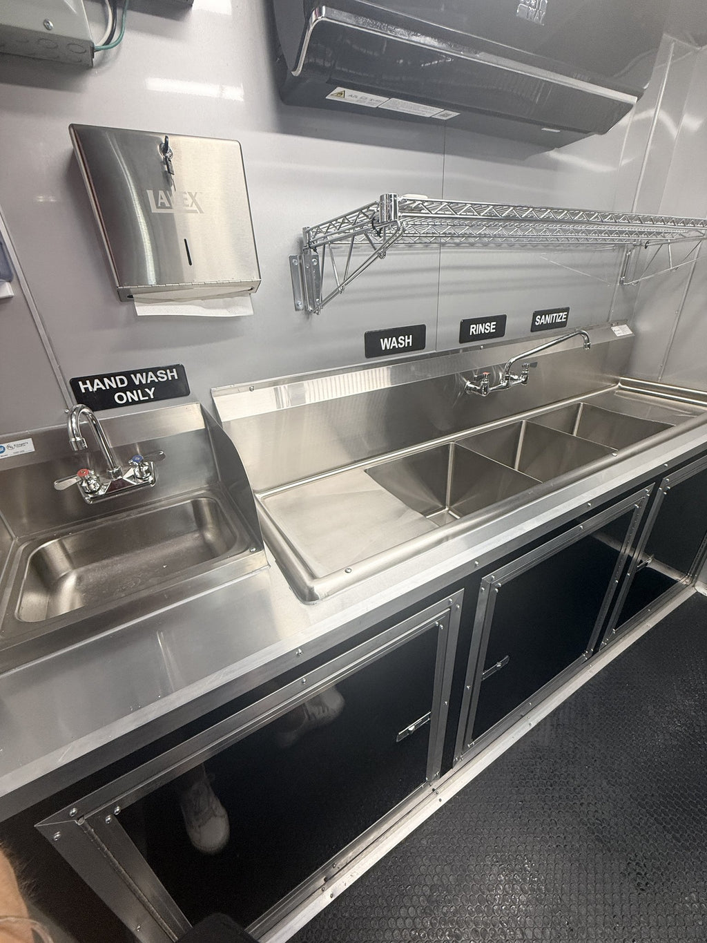 A stainless steel mobile kitchen sink unit with a hand wash sink on the left, a three-compartment rinse and sanitize basin in the center, and a wall-mounted paper towel dispenser above. Black cabinet doors line the base, and labels above the sinks read 'HAND WASH ONLY', 'WASH', 'RINSE', and 'SANITIZE'.