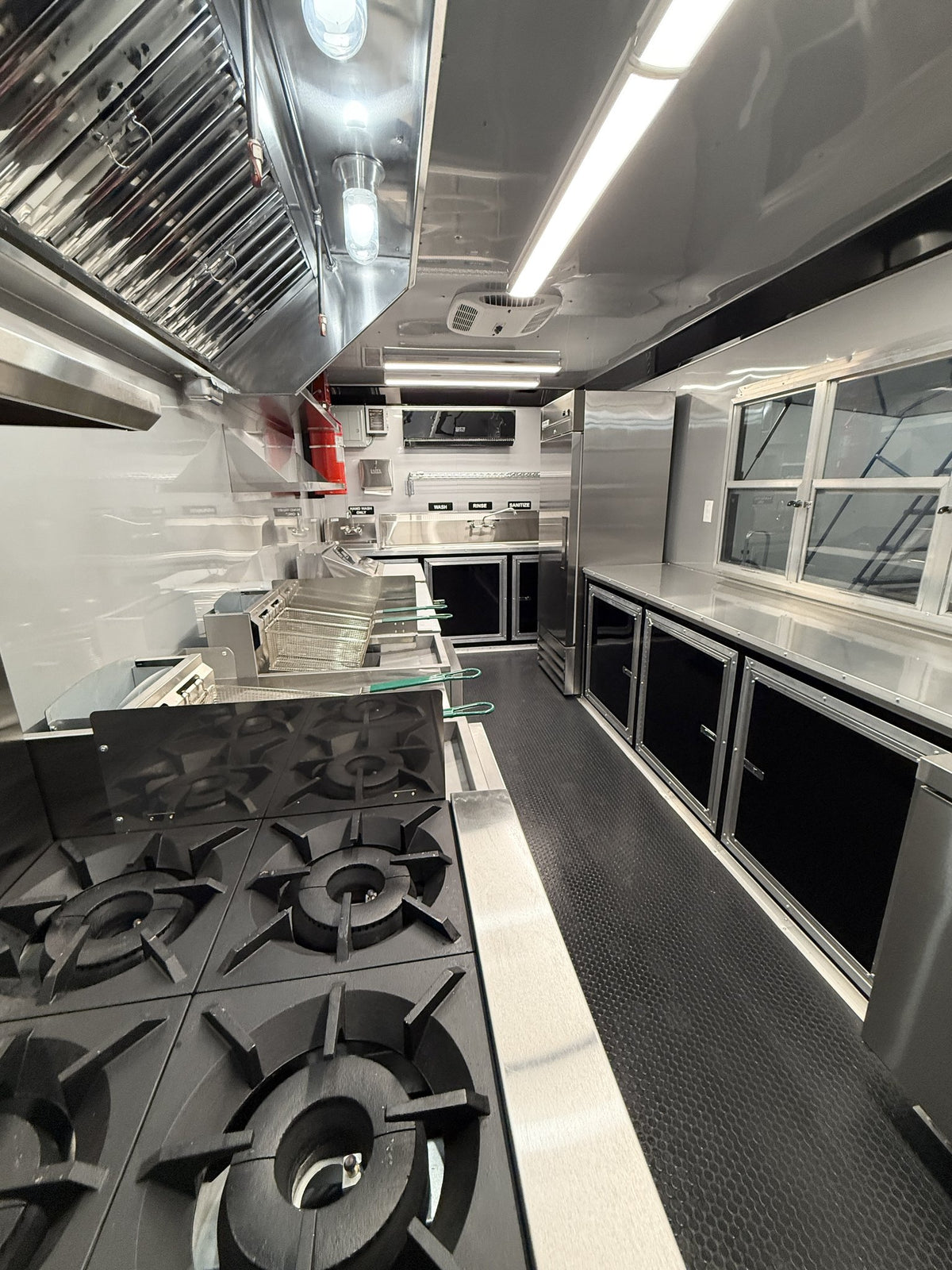 A stainless steel mobile kitchen interior with a gas range in the foreground, prep counters, windows, and overhead ventilation equipment.