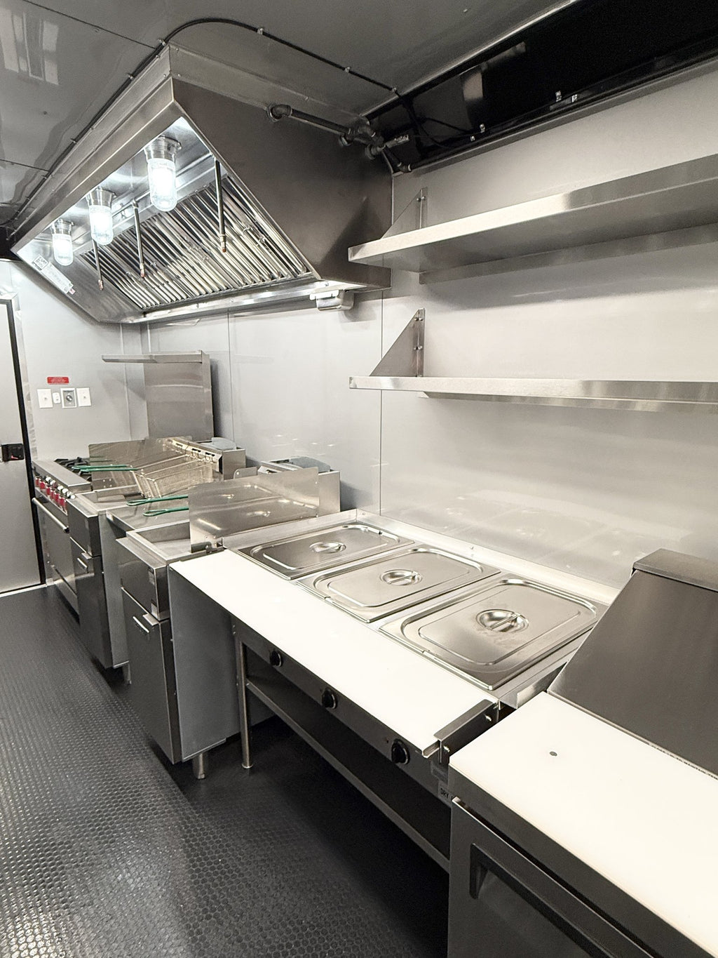A commercial kitchen interior with stainless steel counters, food pan lids, shelving, a large overhead range hood, and stainless steel appliances on a dark floor.