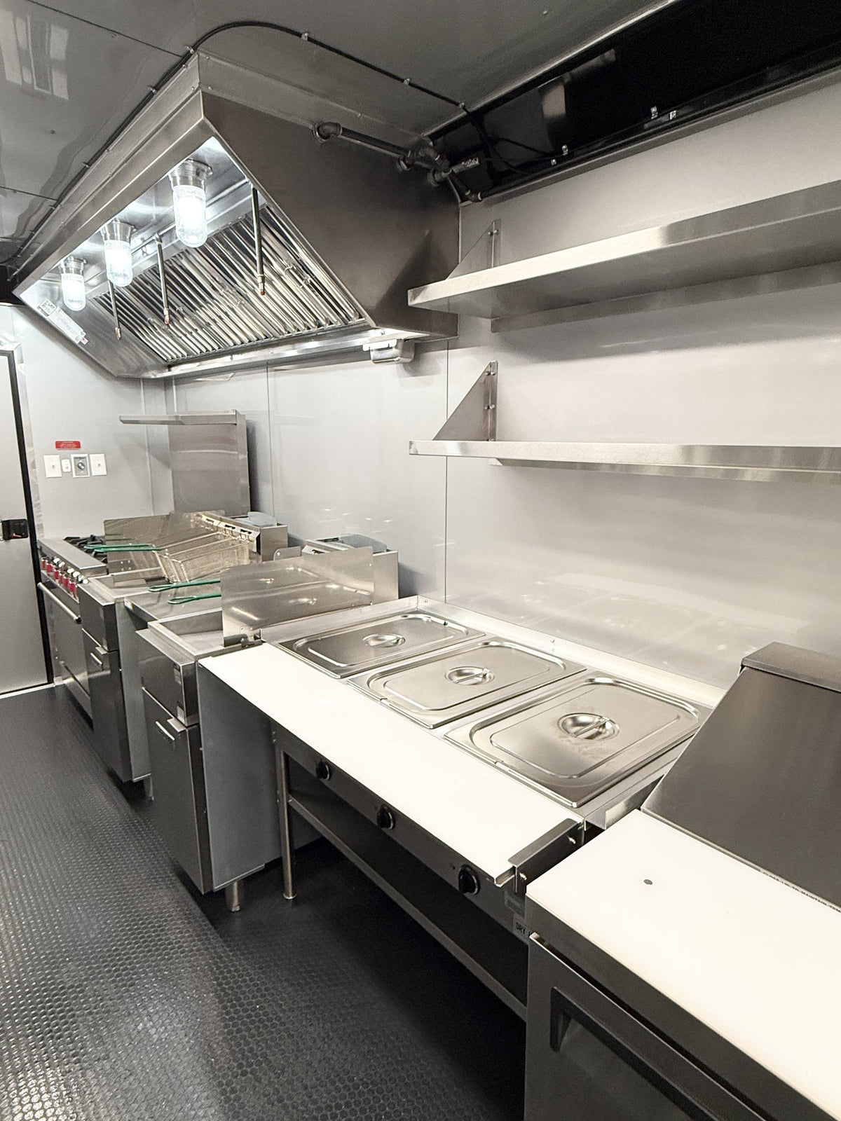 A commercial kitchen interior with stainless steel counters, food pan lids, shelving, a large overhead range hood, and stainless steel appliances on a dark floor.