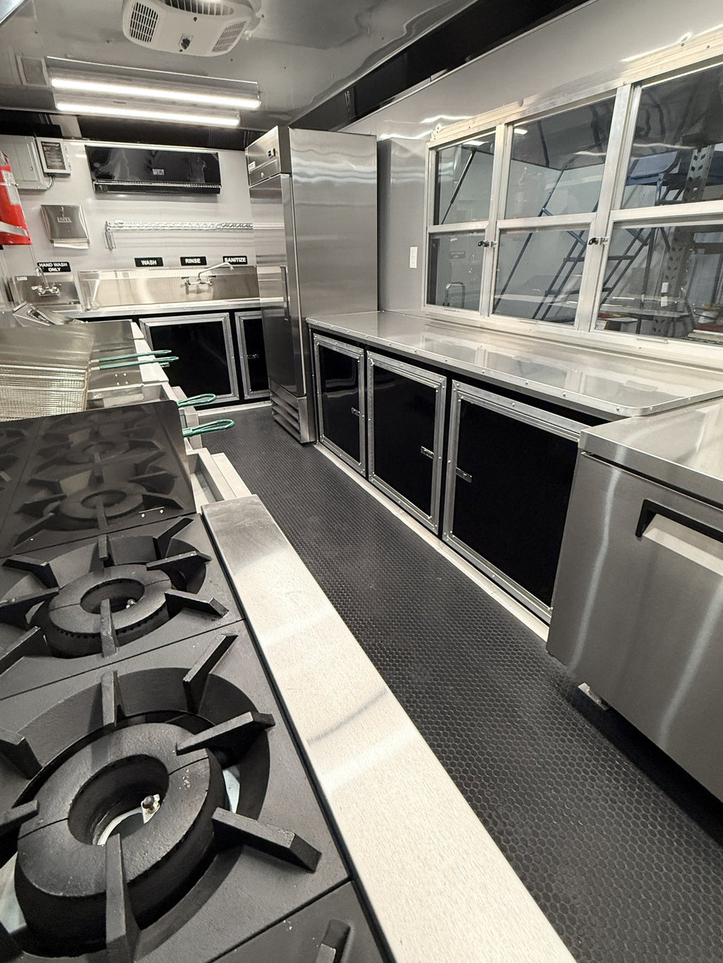 A commercial kitchen interior with stainless steel counters, gas burners, black storage cabinets, and a textured dark floor.