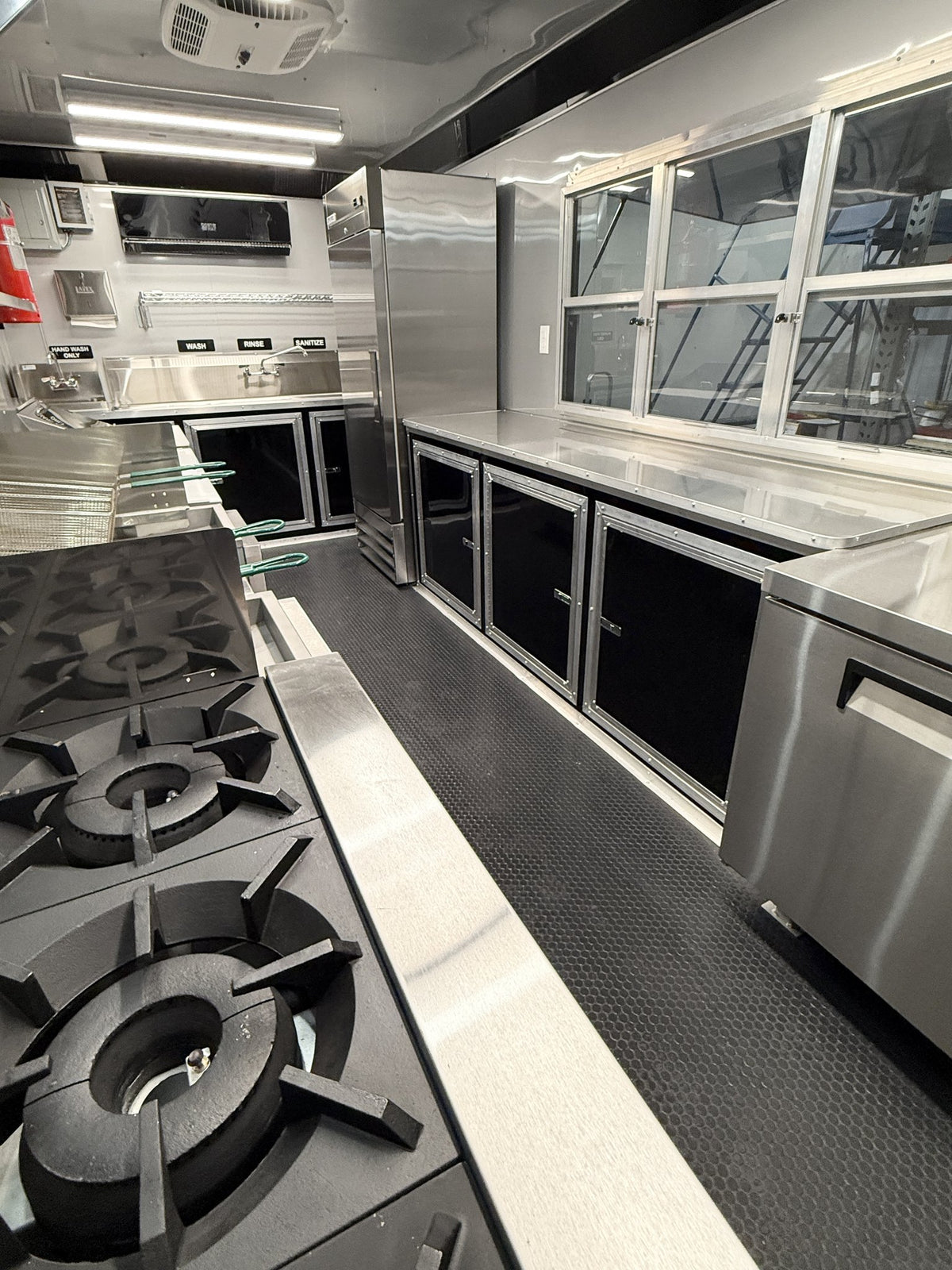 A commercial kitchen interior with stainless steel counters, gas burners, black storage cabinets, and a textured dark floor.