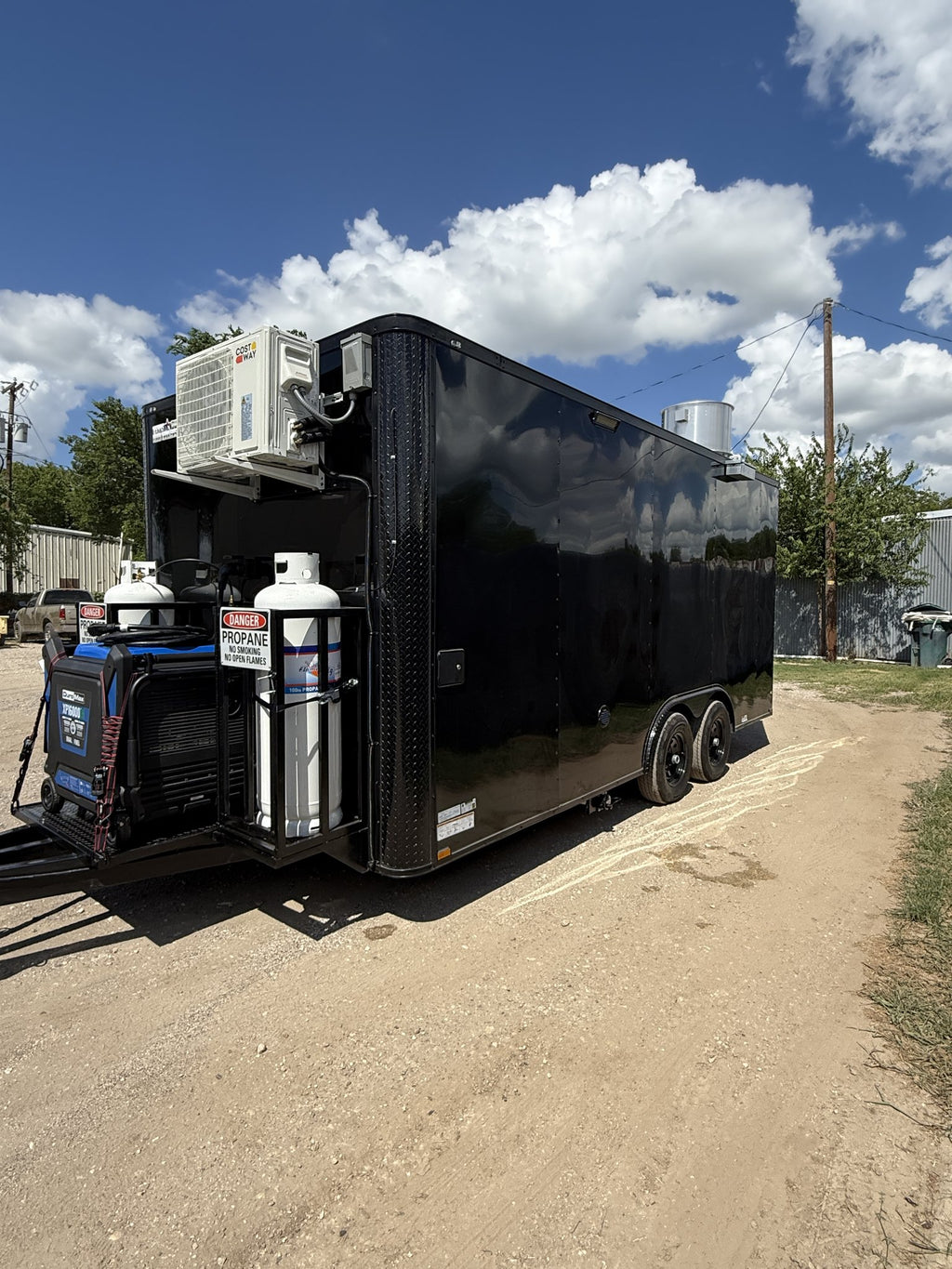 A black enclosed utility trailer parked outdoors on a dirt road under a blue sky with clouds. The trailer is equipped with an air conditioning unit, multiple white propane tanks, and a blue generator mounted on the rear hitch area.