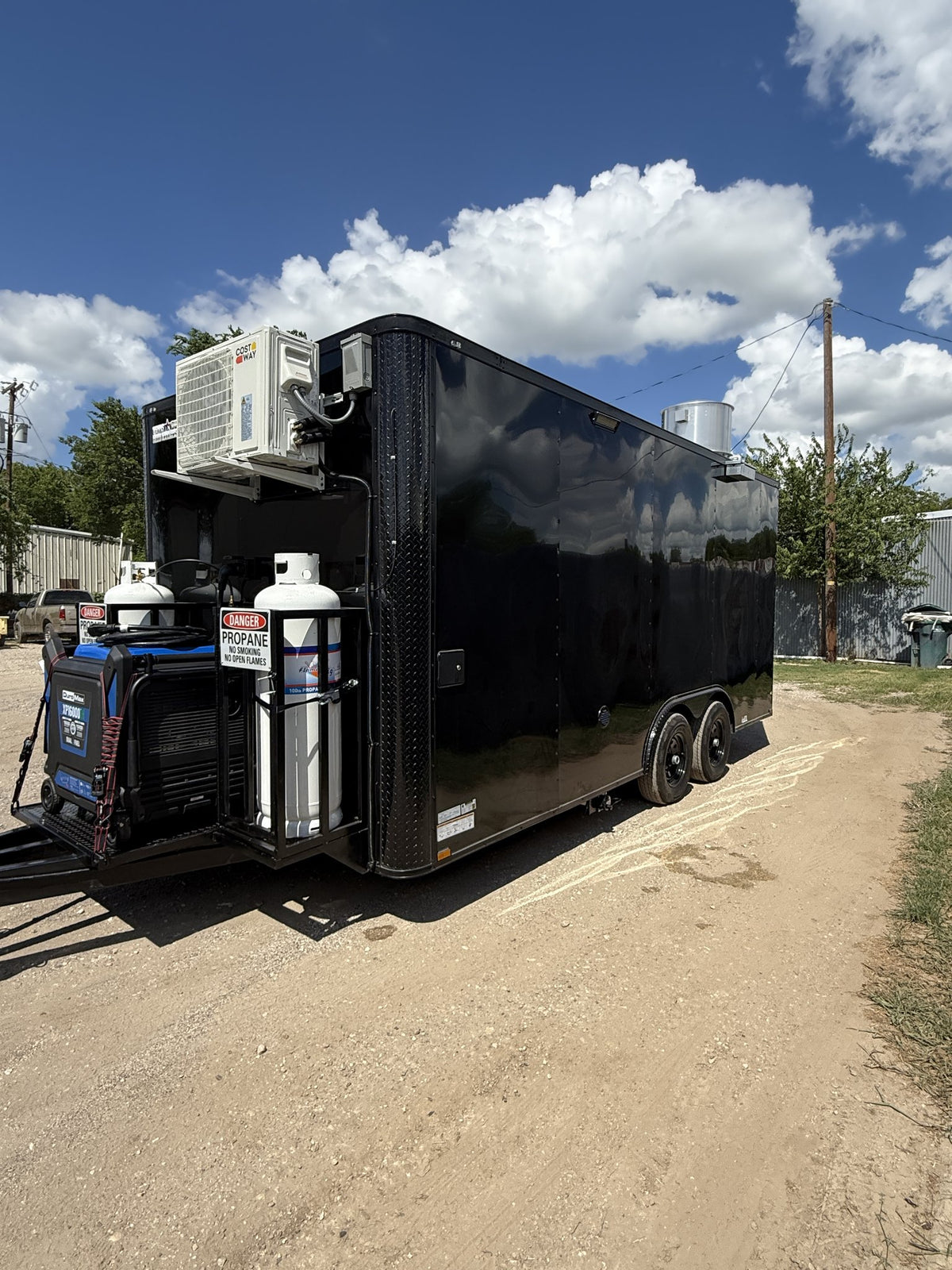 A black enclosed utility trailer parked outdoors on a dirt road under a blue sky with clouds. The trailer is equipped with an air conditioning unit, multiple white propane tanks, and a blue generator mounted on the rear hitch area.