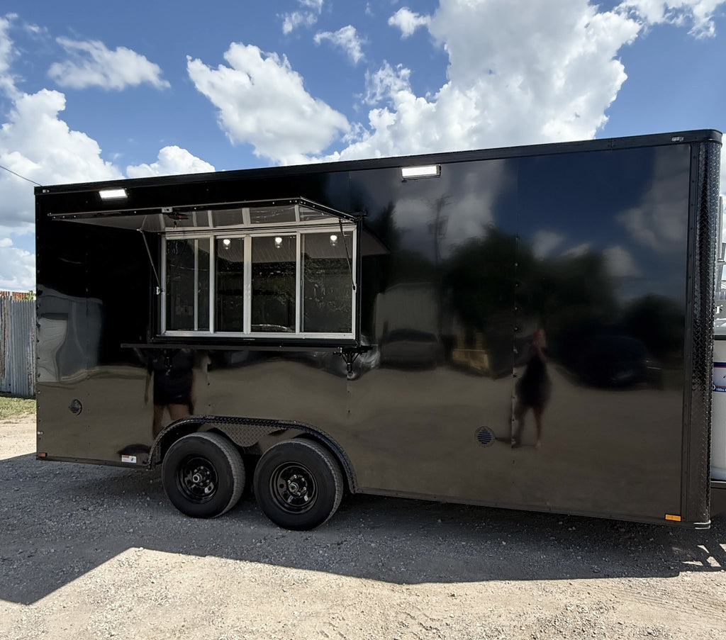 A black enclosed food truck trailer parked outdoors on gravel under a blue sky with clouds. The side of the trailer has a large service window with an extended awning, and the glossy black exterior reflects the photographer and surrounding environment.