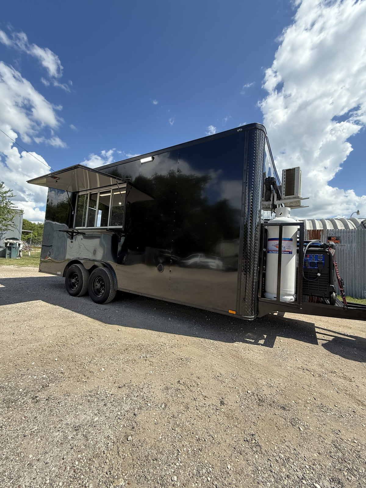 A black food truck trailer parked outdoors on a gravel lot under a blue sky with clouds. The side of the trailer has an open serving window and a small awning, with a large white water tank mounted on the rear right side.